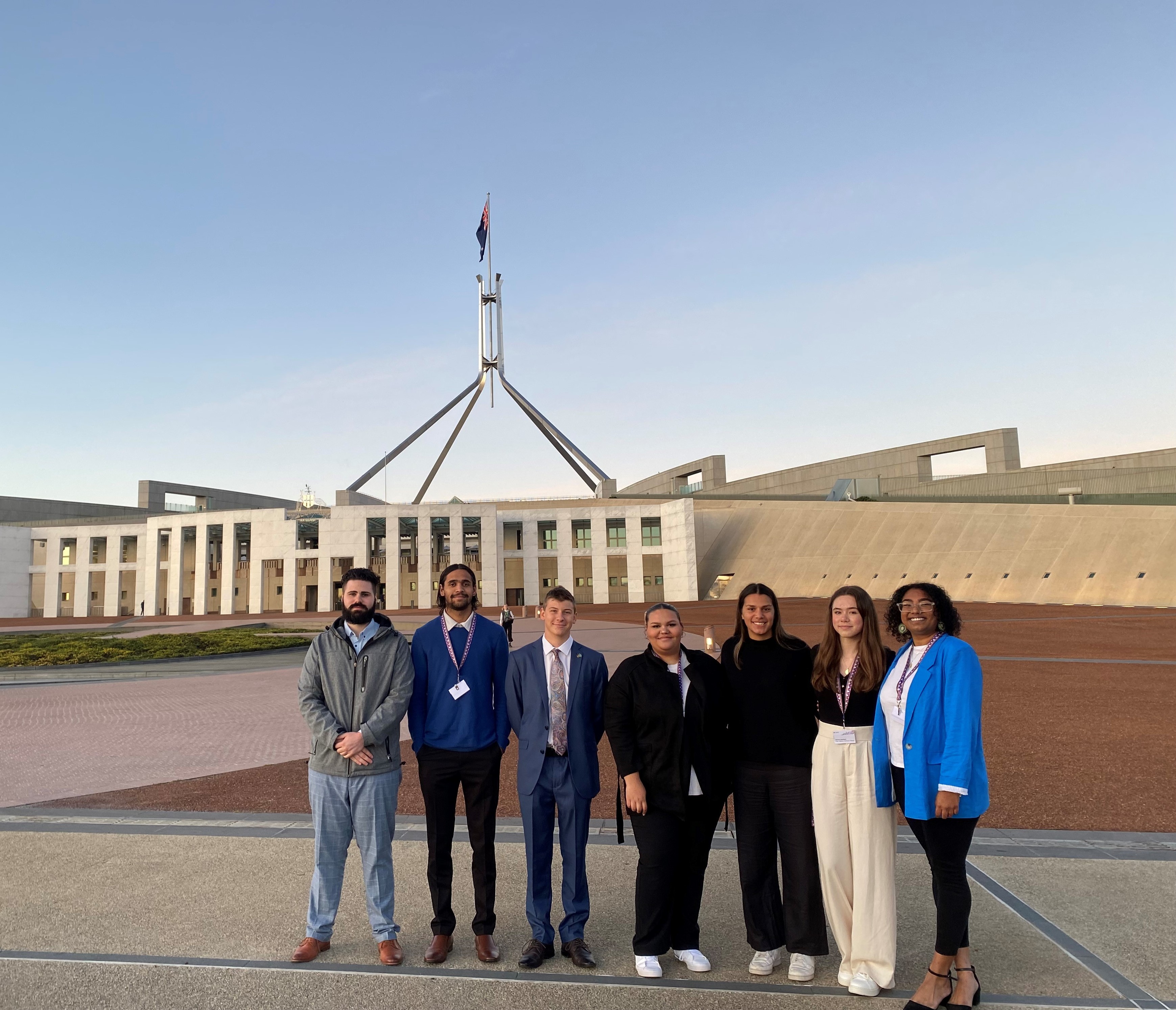 Three young men and four young women representing first nations people standing in front of Australian parliament house.