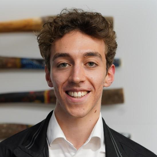 Head shot of Brodie. Young person smiling with short brown curly hair wearing a black jacket and white collared shirt.