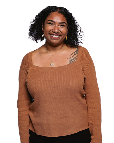 Rhea headshot - person smiling with dark curly hair wearing a long sleeved brown top.