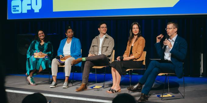 5 forum panel members sitting on seats on a stage listening to one of the panel members speaks