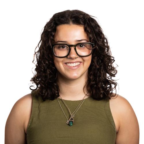 Image of Lottie smiling with shoulder length curly hair, wearing spectacles, a sleeveless top and a necklace with a pendant.