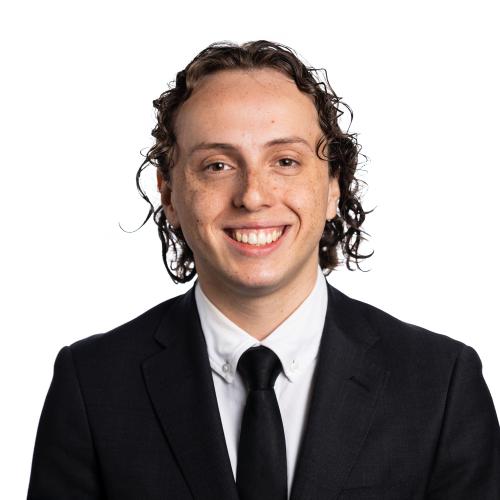 Image of Ramon smiling with medium length curly hair, wearing a collared shirt, tie and a blazer.