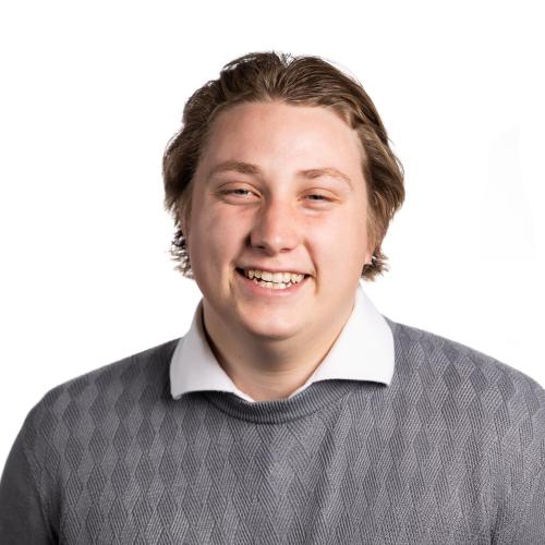 Headshot of Reede smiling at the camera with short hair wearing a collared shirt and a knitted jumper.